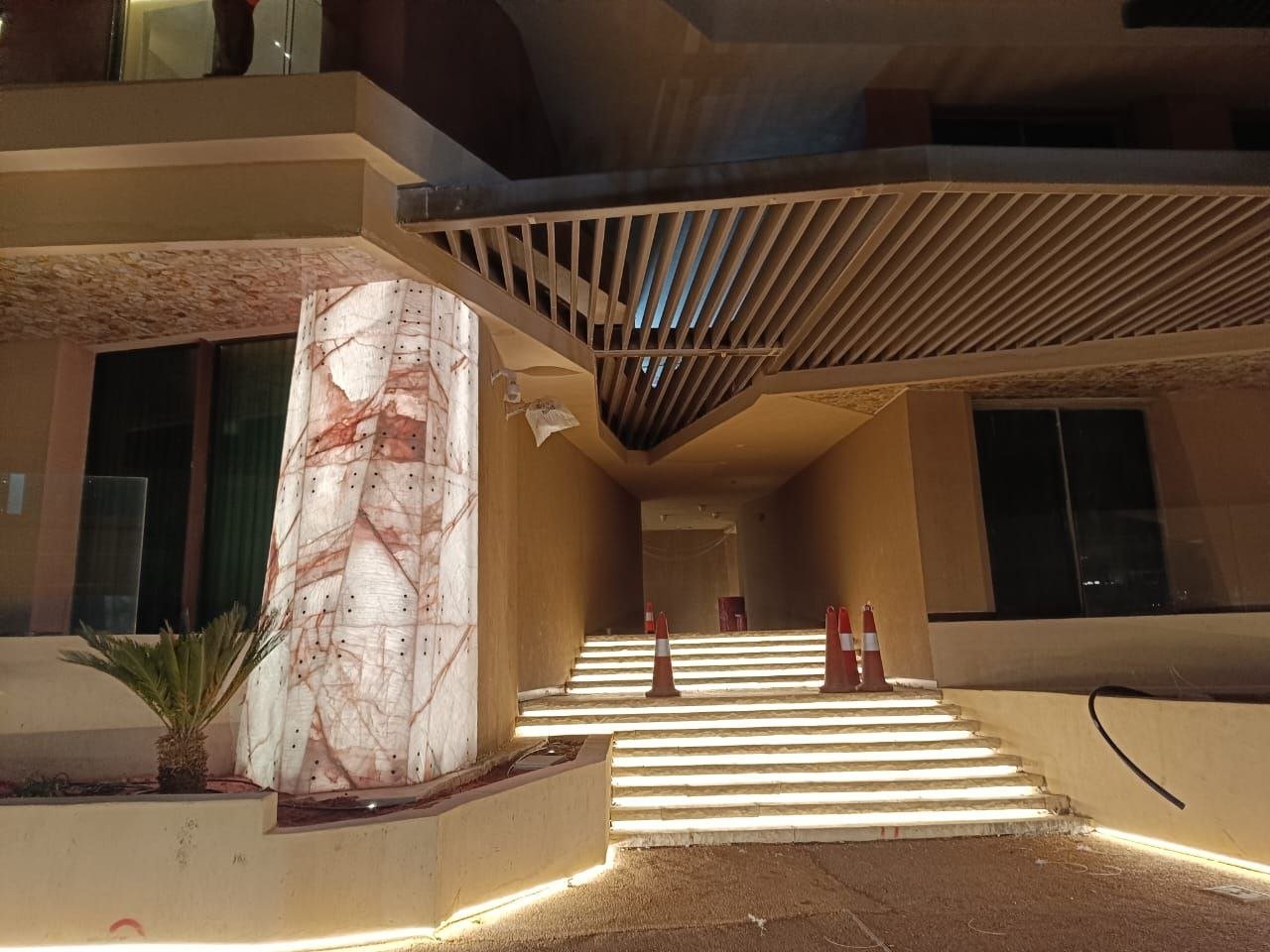 A NEOM Sindalah building entrance at night: a back-lit marble pillar glowing from within, illuminated stair treads, slatted ceiling above.