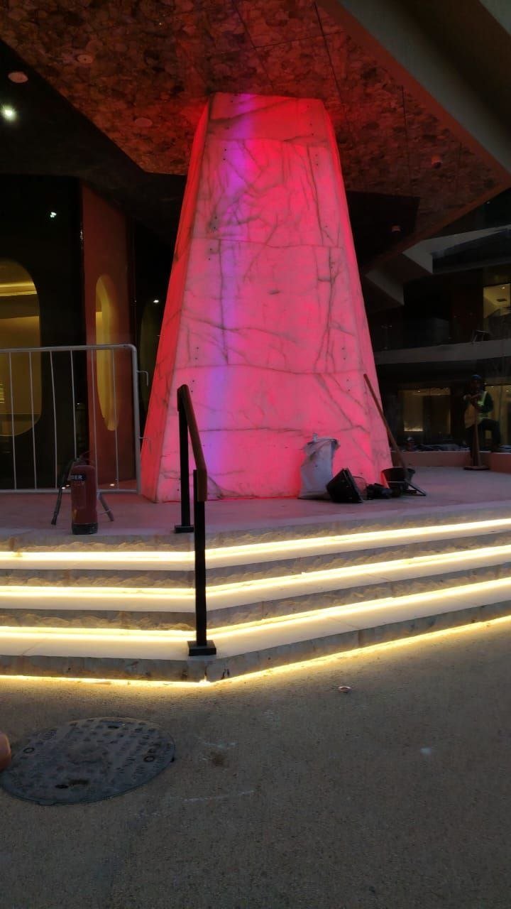 A tall marble-clad obelisk lit in deep pink and red from the inside, with warm yellow LED strips wrapping the stair base.